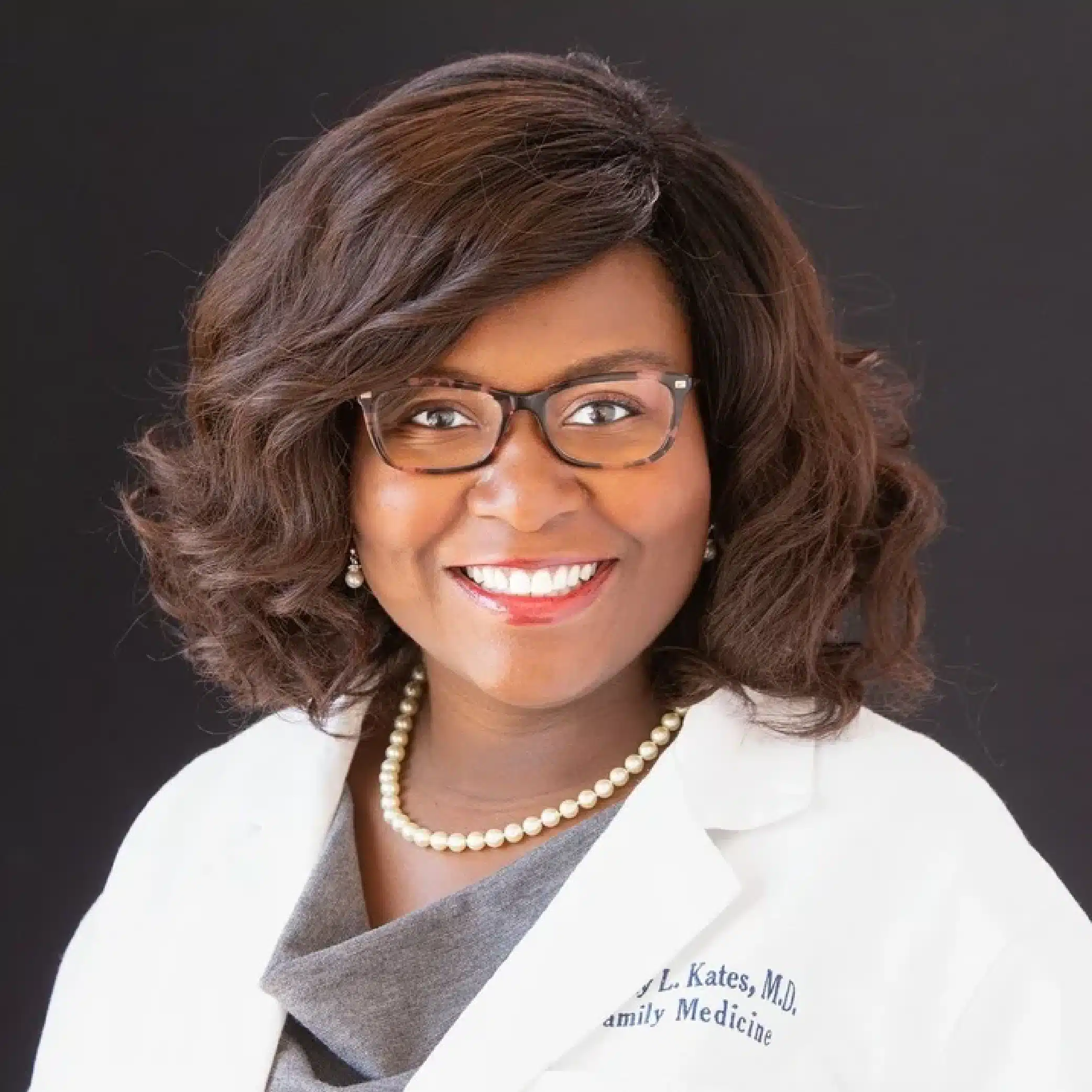 Portrait of a smiling African American female doctor in a white coat with a pearl necklace against a dark background.