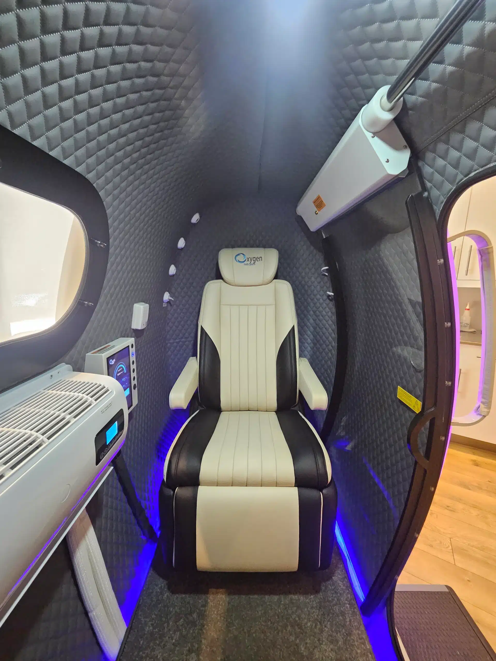Interior of a compact medical capsule with quilted grey walls and a beige black seat labeled Oxygen, blue LED floor lighting, window on left, and equipment mounted along the wall.