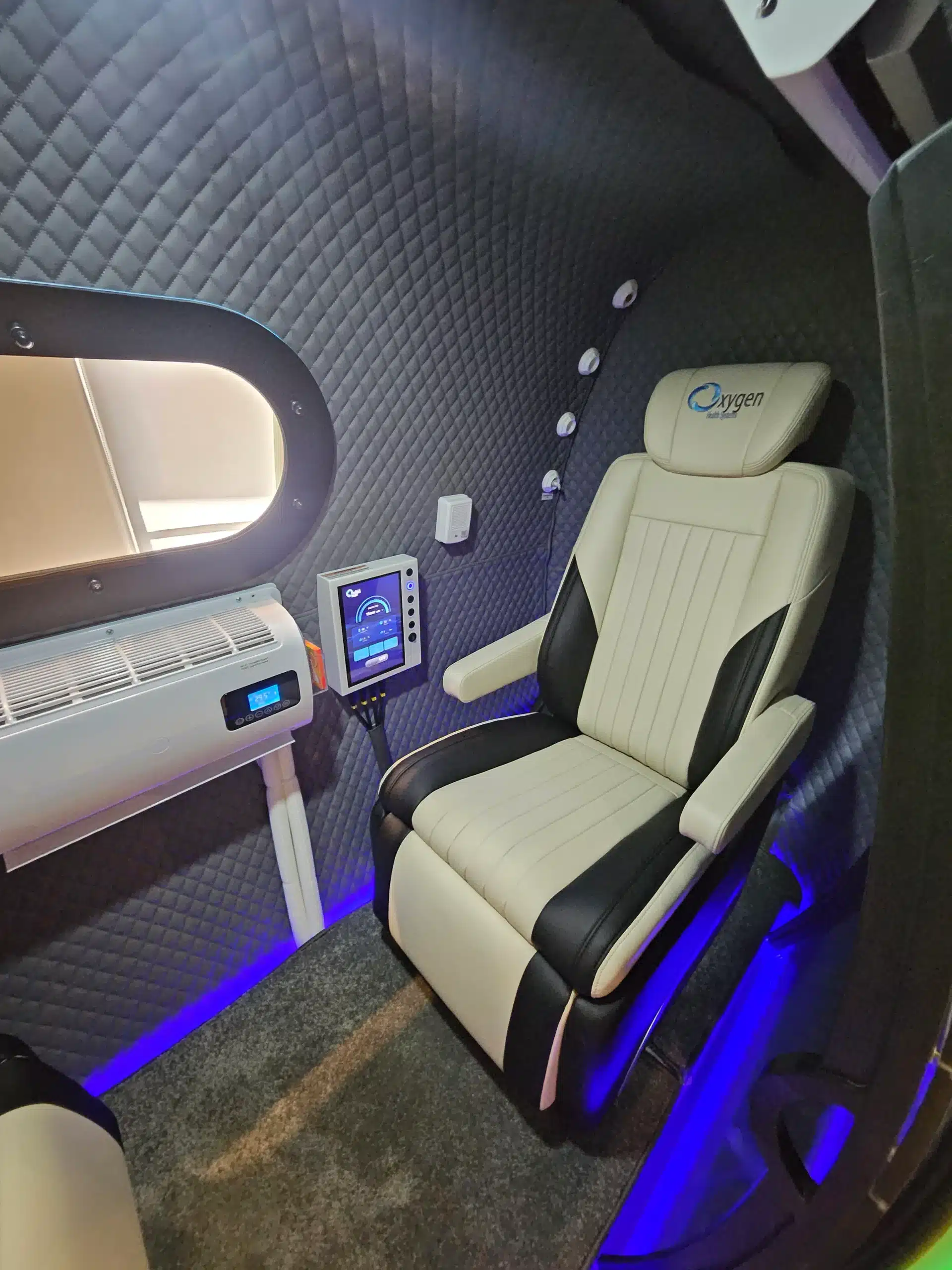 Luxurious pod interior with quilted dark walls, cream leather captain chair, medical/controls panel, and blue ambient lighting near a window.